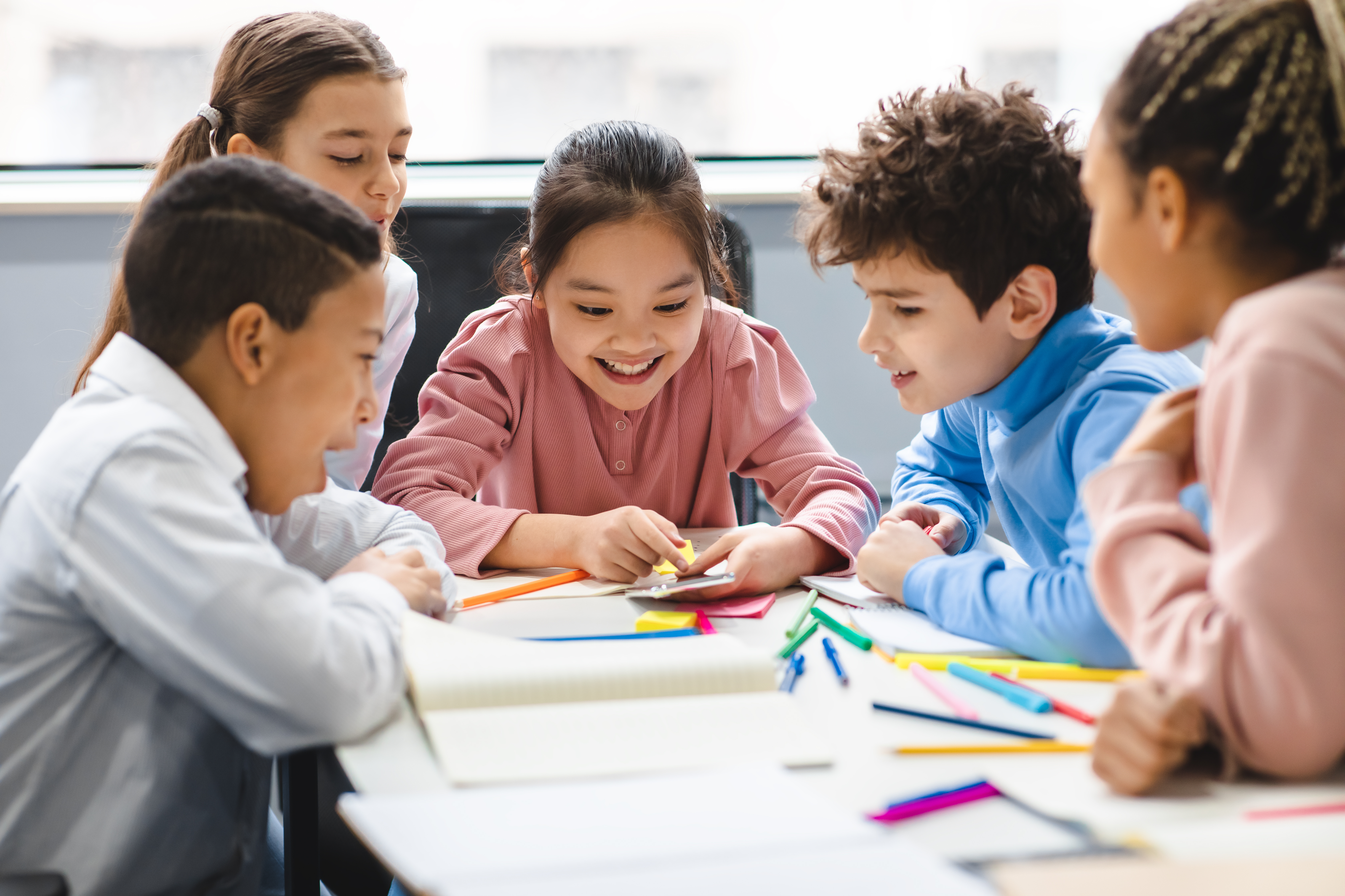 Technology And Pupils Concept. Group of excited multicultural happy junior children sitting at table and using smartphone, playing online mobile games. Modern Device, Gadget Addiction