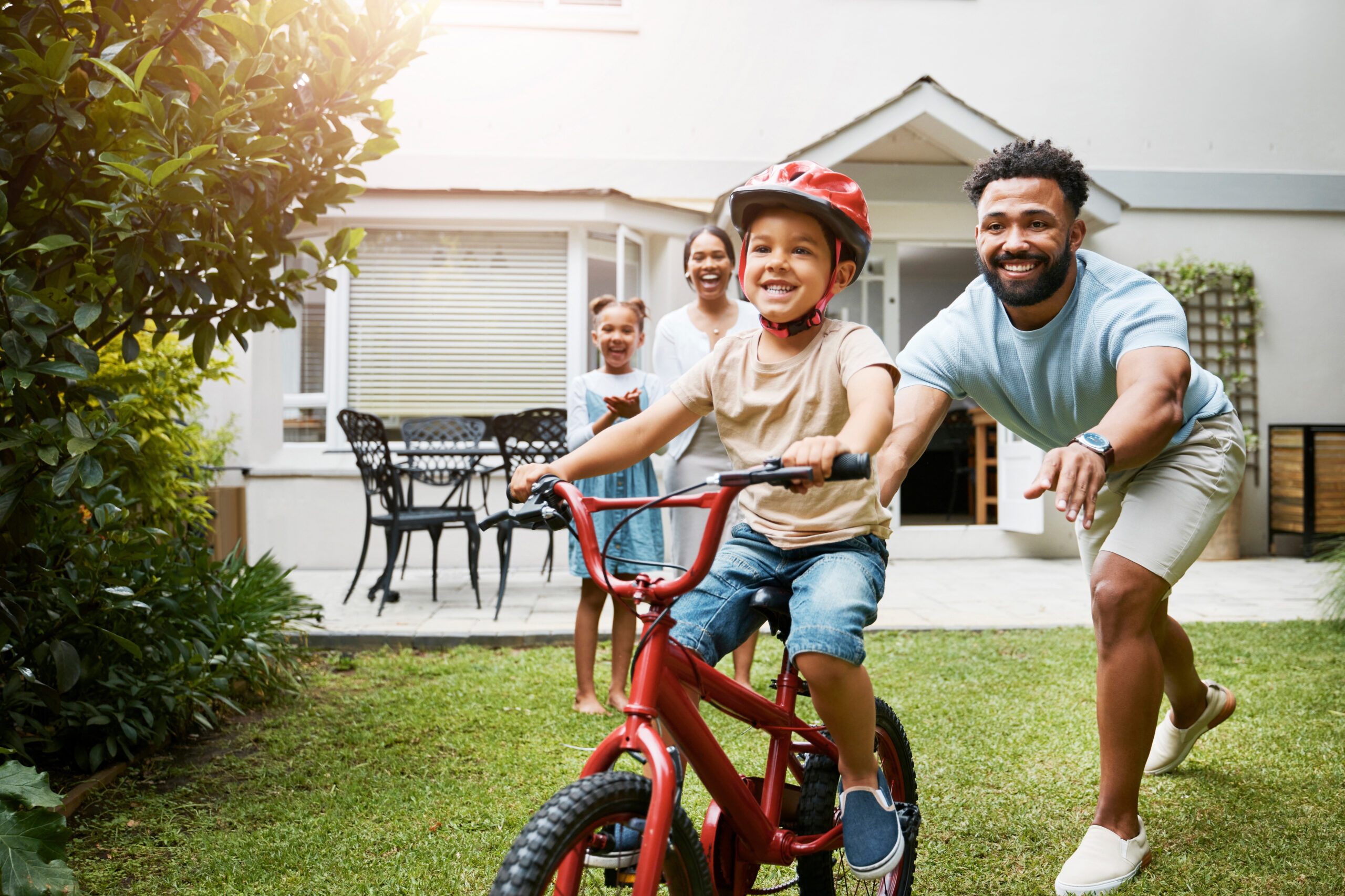 Learning, bicycle and proud dad teaching his young son to ride while wearing a helmet for safety in their family home garden. Active father helping and supporting his child while cycling outside.