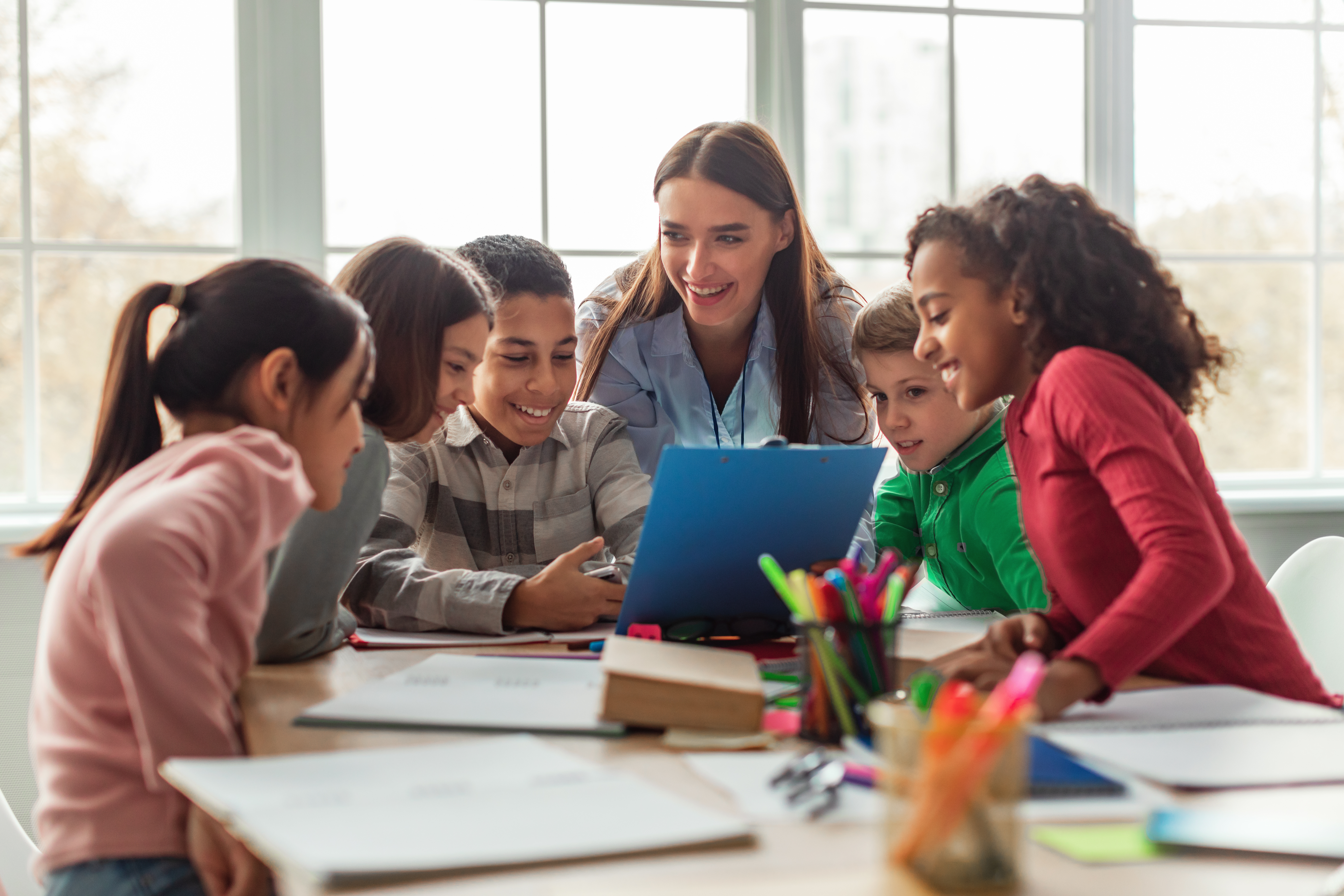 Happy Diverse School Children And Teacher Woman Having Class Sitting At Desk In Classroom At School. Modern Education And Knowledge Concept. Selective Focus