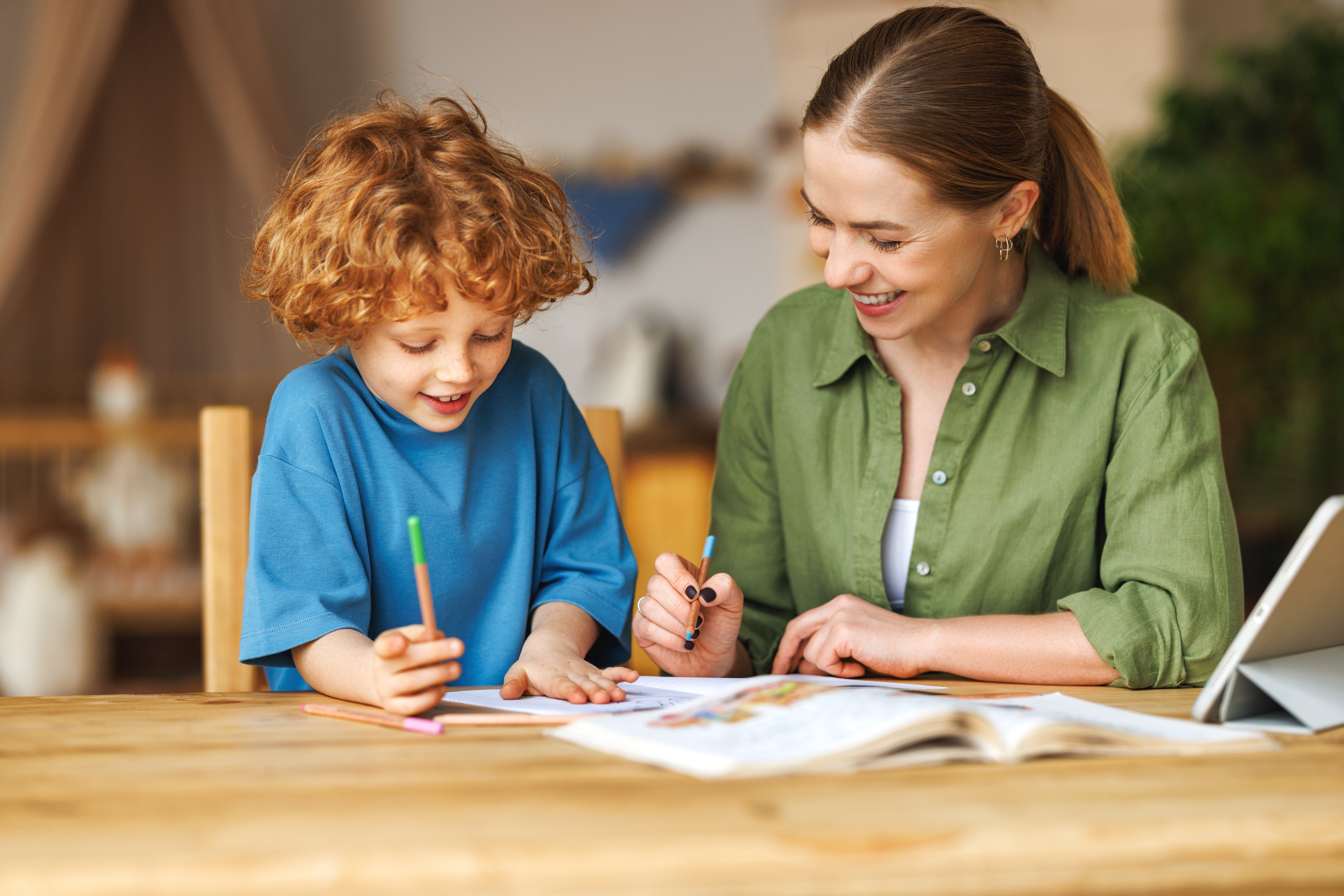 Smiling teacher or mother and little schoolboy doing exercises in copybook while studying remotely via tablet,   while doing assignment together and studying remotely at home