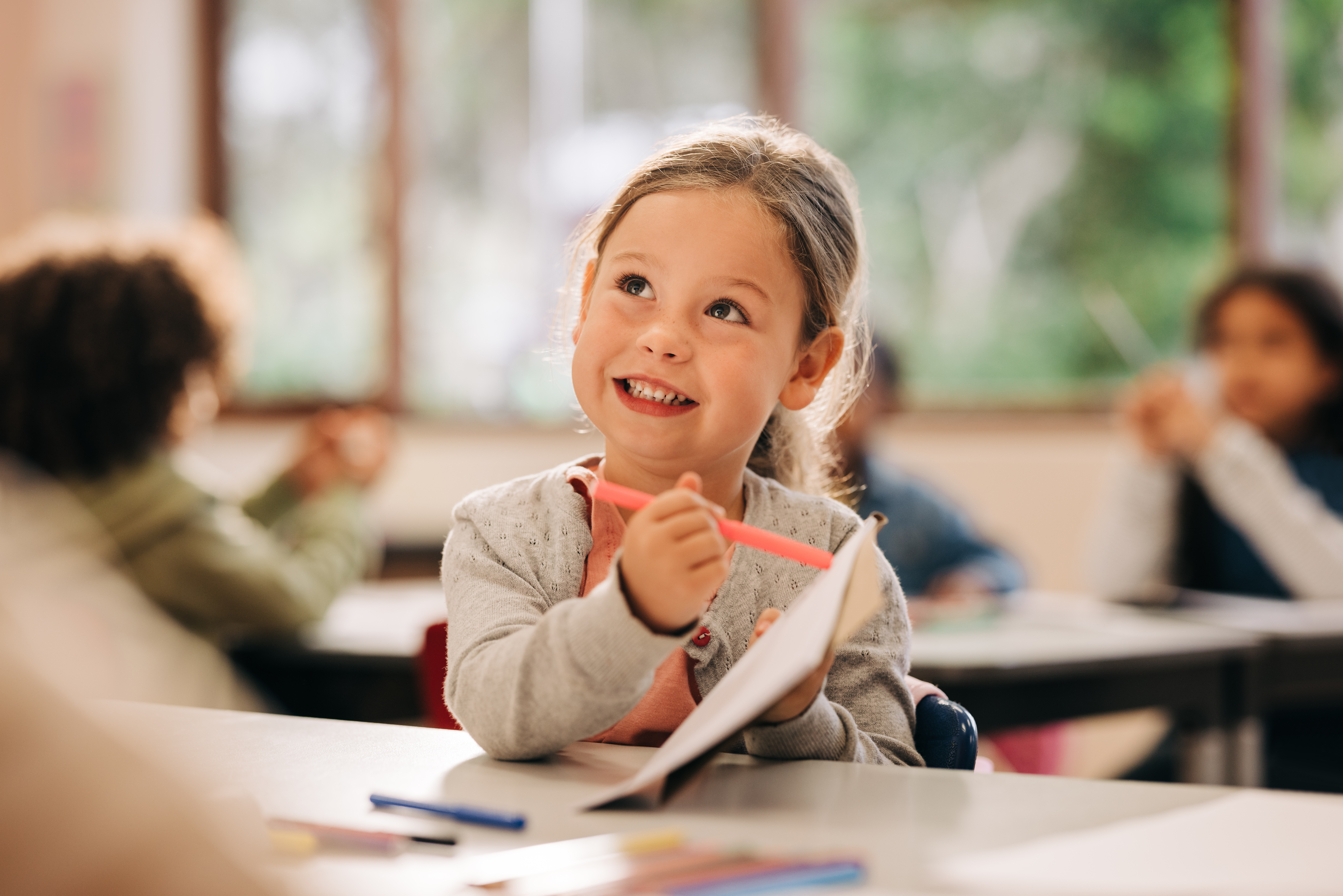 Excited little girl learns to draw with a colour pencil in an art class. Happy elementary school kid talks to her teacher while holding a colour pen and a drawing book. Creativity as a part of primary education.