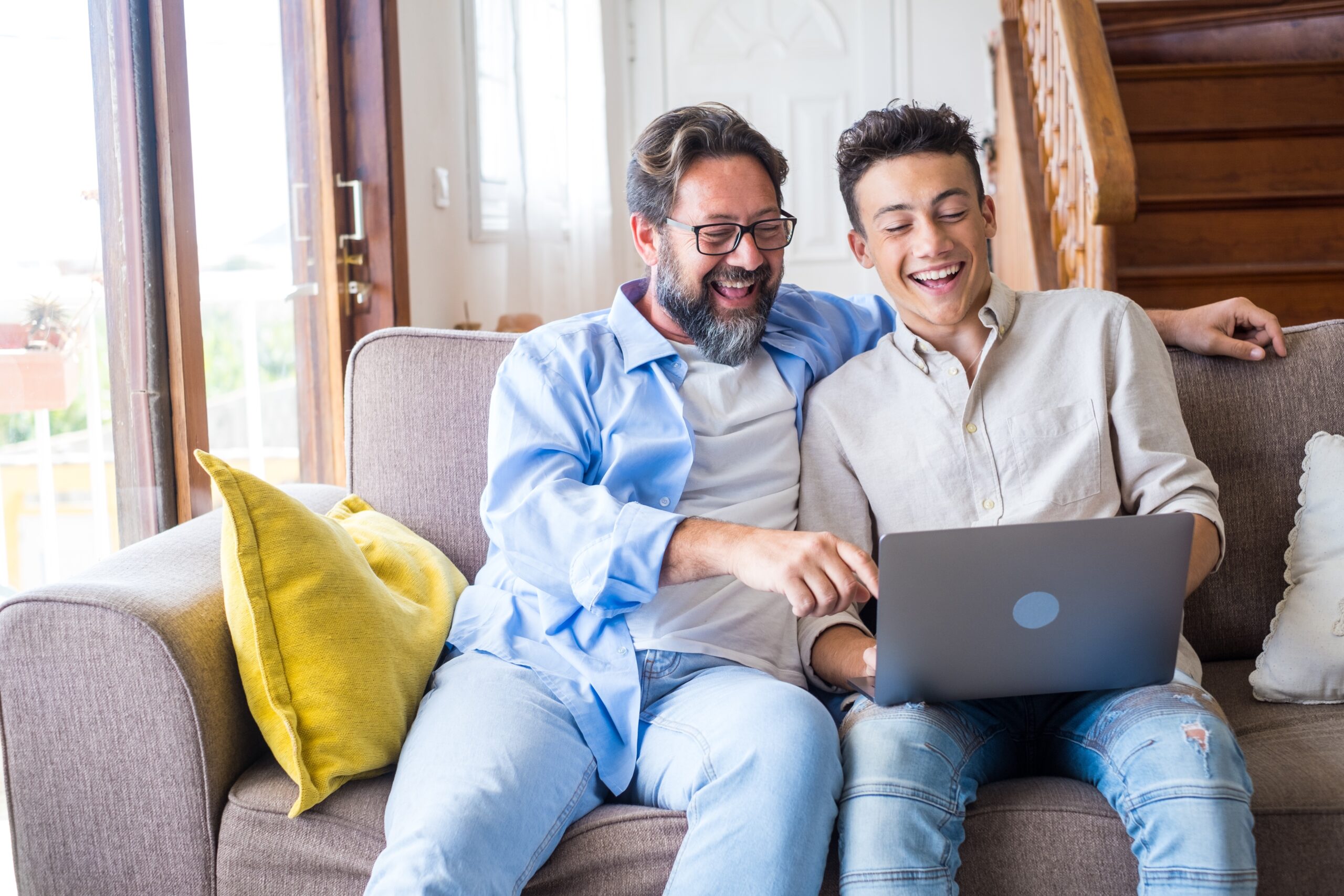 Happy father and son looking at laptop screen while sitting on couch in living room at home. Father with teenage son browsing social media content using laptop in living room